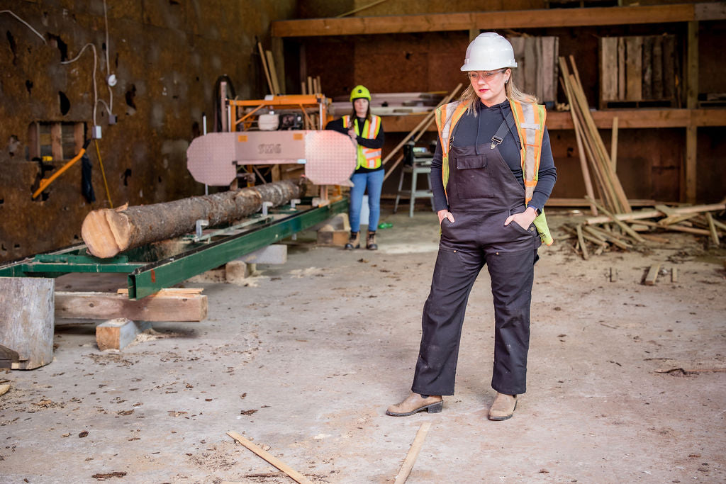 Woman wearing high-quality workwear overalls designed specifically for women, and a construction vest, with lumber site in the background.